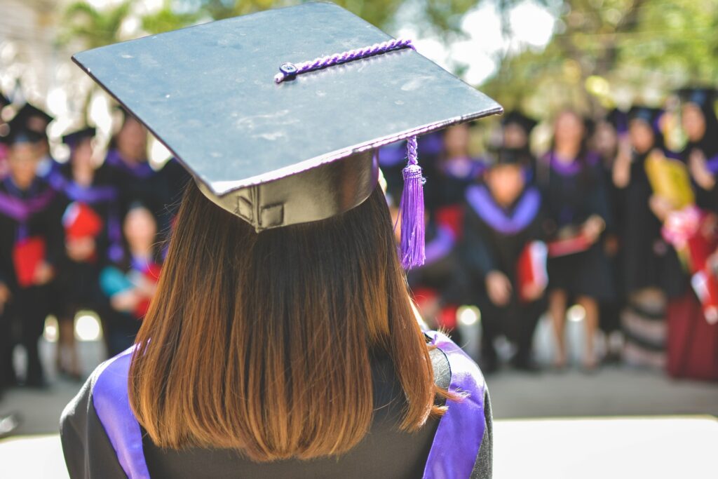 From the back, a graduate wearing cap and gown, blurred graduates in the background