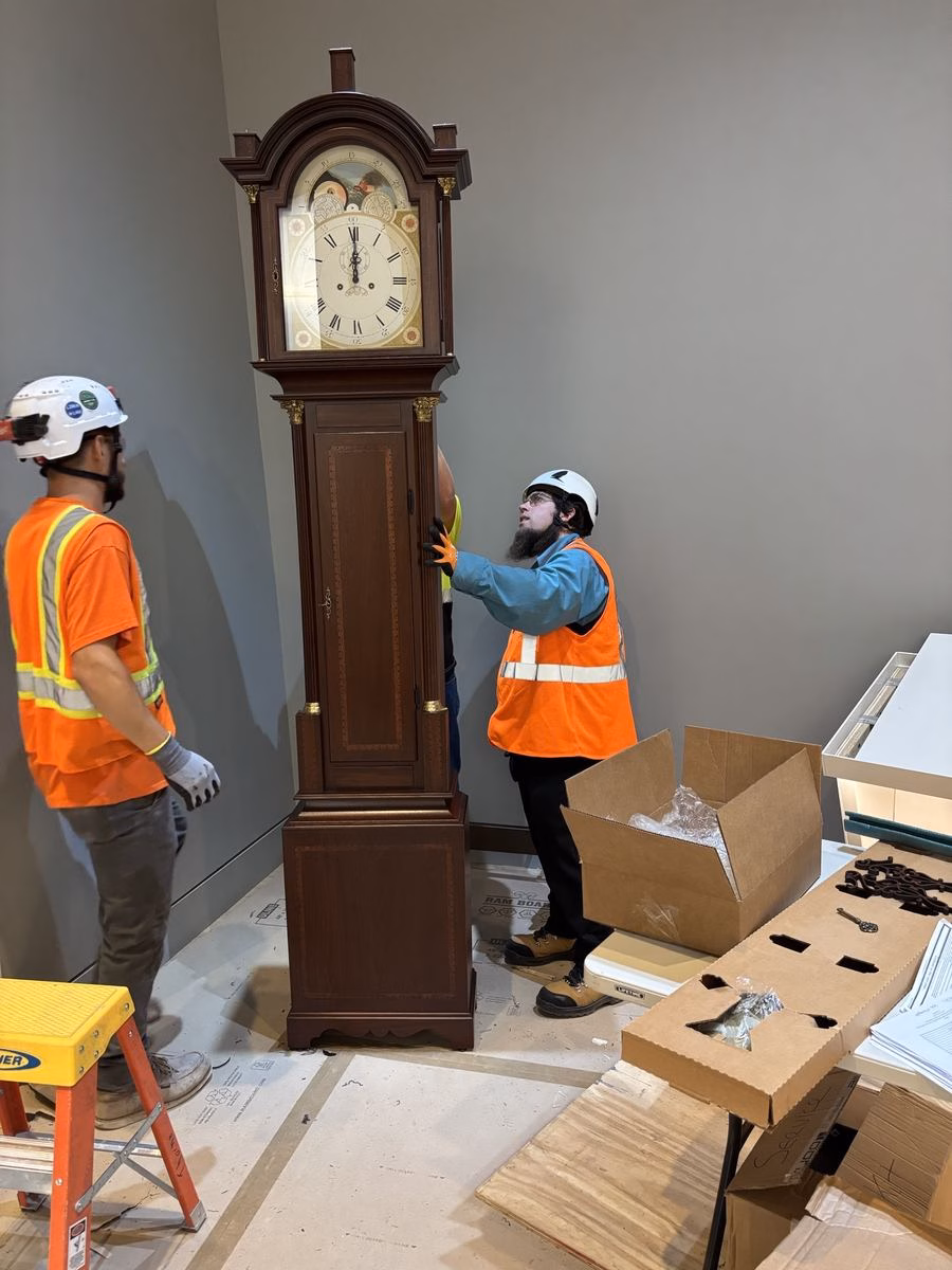 Craftsmen in hard hats and safety vests setting the finished grandfather clock replica in place at the Obama Presidential Center